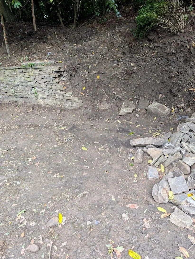 A damaged stone wall with a pile of rocks and dirt in a wooded area.